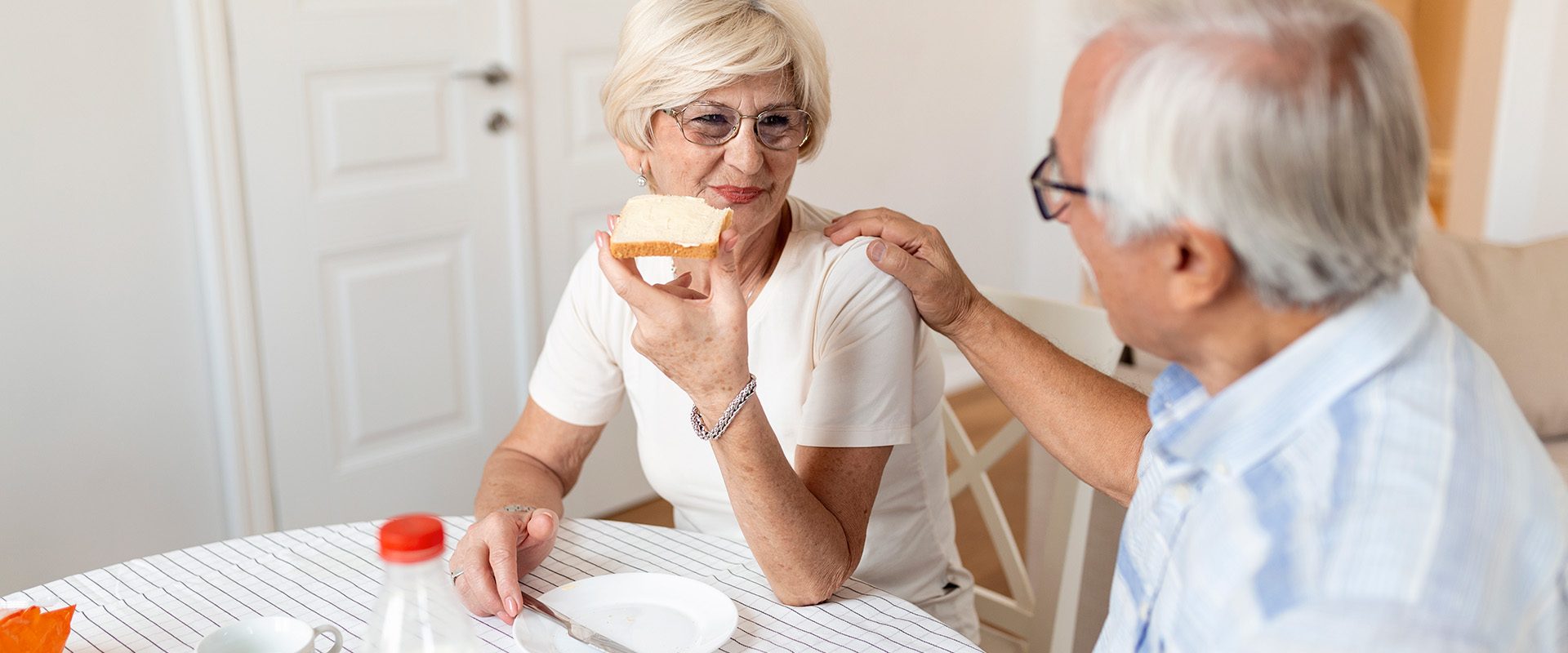 cheerful-senior-couple-having-breakfast-home-elderly-man-hugging-woman-mature-woman-holding-piece-bread-old-man-woman-sitting-table-talking