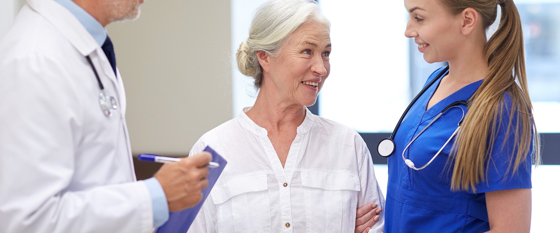 medicine-age-health-care-people-concept-male-doctor-with-clipboard-young-nurse-senior-woman-patient-talking-hospital-corridor